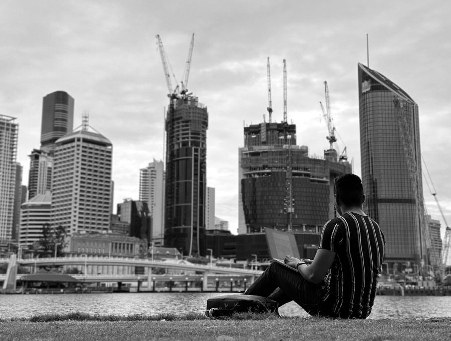 Zach sitting on the riverbank with a laptop, facing the Brisbane skyline and its construction cranes.
